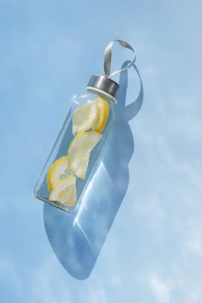 Glass bottle with lemon water drink detox at sunlight on abstract blue water texture, copy space. Healthy natural infused water is boost metabolism, weight loss. Aesthetic still life, refreshing drink