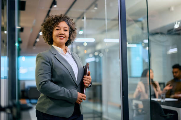 Portrait of confident insurance agent in the office looking at camera. Happy financial consultant at work looking at camera. Her clients are in the office in the background. bank manager stock pictures, royalty-free photos & images