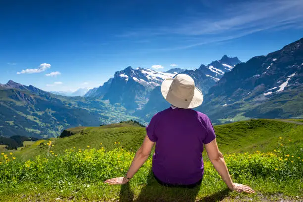 Senior Korean woman on a trail from Kleine Scheidegg to Mannlichen with the Eiger in the background and Snowcapped Swiss Alps - taking the time to sit amongst the wildflowers and take in the fabulous views in the Swiss Alps