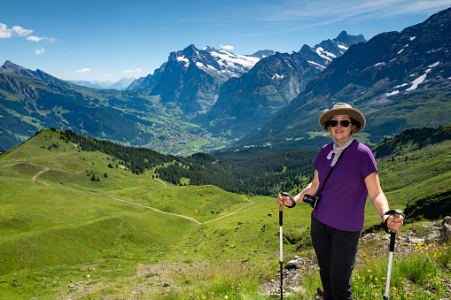 Mature Korean woman hiking the Mannlichen trail to Kleine Scheidegg, stops to pose for a portrait with the town of Grindelwald and the mounatins of First, Wetterhorn and Schreckhorn in the background.