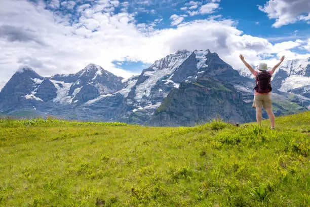 Senior Asian woman, with arms extended, enjoying the majestic views when hiking through the Mountain view trail in the Swiss Alps in the Jungfrau region. The Mountain view trail hike from Murren and leads over the Alpine meadows and through forests from Allmendbhubel to Grutschlap with fantastic views of Eiger, Monch and Jungfrau peaks.