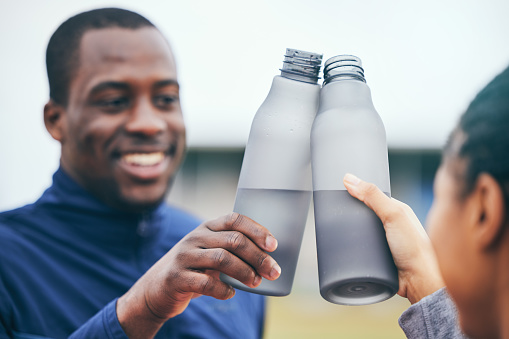 Fitness, water bottle and black couple toast outdoors together after workout, exercise or training. Sports targets, celebration and man and woman cheers with liquid for hydration to celebrate goals.