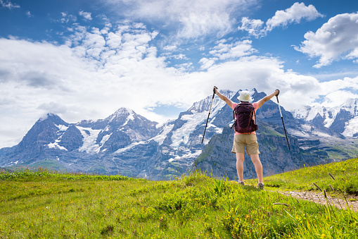 Senior Asian woman, with arms extended, enjoying the majestic views when hiking through the Mountain view trail in the Swiss Alps in the Jungfrau region. The Mountain view trail hike from Murren and leads over the Alps meadows and through forests from Allmendbhubel to Grutschlap with fantastic views of Eiger, Monch and Jungfrau peaks.