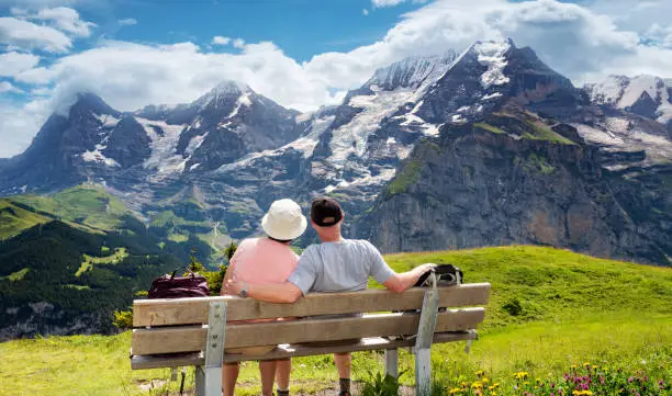 Senior couple enjoy view of Eiger and Jungfrau alps from a rest area for hikers on the mountain view trail in the Jungfrau region of the Swiss alps. The Mountain view trail hike from Murren and leads over the Alps meadows and through forests from Allmendbhubel to Grutschlap with fantastic views of Eiger, Monch and Jungfrau peaks.