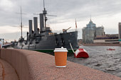 paper cup of natural coffee on bridge of river next to ship cruiser Aurora in St. Petersburg, Russia
