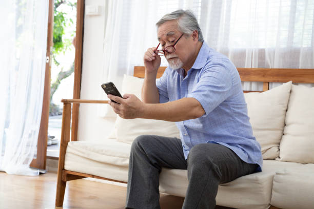 senior asian man moves his reading glasses to look at mobile phone while sitting on the living room couch at home, eye problems concept - macular degeneration imagens e fotografias de stock