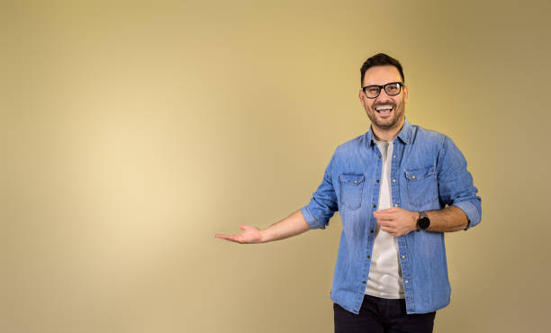 portrait of cheerful male entrepreneur gesturing towards copy space while standing isolated on beige background. happy businessman laughing and doing advertisement while looking at camera - boegbeeld model stockfoto's en -beelden