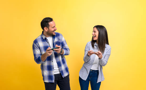 istock Young couple laughing cheerfully and playing video game over smart phones while standing against yellow background. Excited man and woman dressed in casuals competing and looking at each other 1483271313