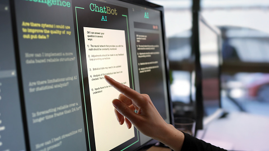 Close-up stock photograph showing a touchscreen monitor being used in an open plan office.
A woman’s hand is asking an AI chatbot pre-typed questions & the Artificial Intelligence website is answering.