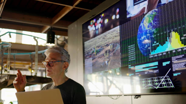 Mature man energy lecture A Stock photograph of a man conducting a seminar / lecture with the aid of a large screen. The screen is displaying data & designs concerning low carbon electricity production with solar panels & wind turbines. These are juxtaposed with images of conventional fossil fuel oil production. climate stock pictures, royalty-free photos & images