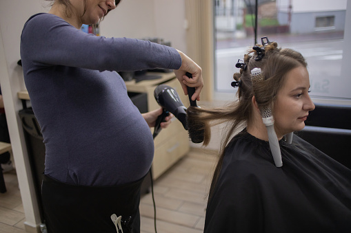 Young pregnant woman, female hairdresser, is working in beauty salon. She is drying her customer's hair.