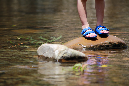 The Asian baby boy is standing on a stone in the stream wearing slippers