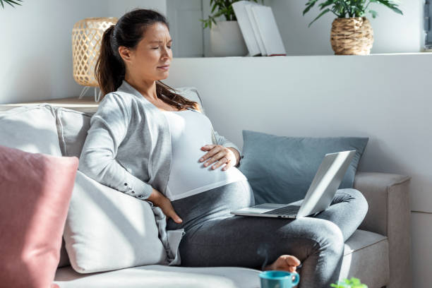 donna incinta preoccupata con mal di stomaco che lavora con il computer portatile seduta sul divano nel soggiorno a casa - cramping foto e immagini stock