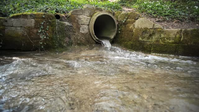Small water stream running out of an industrial-size concrete pipe in a forest in Europe. Daytime, slow motion, no people, wide angle