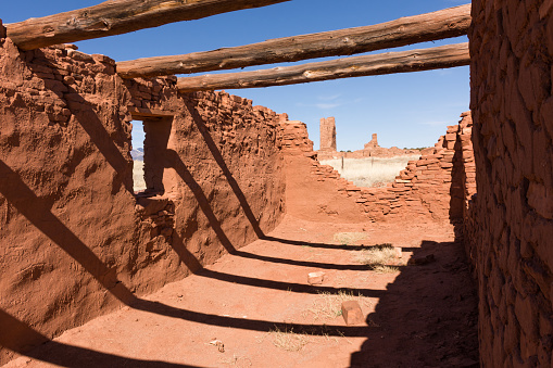 Heavy roof beams, shadows, and window of small mission at Abo Ruins. Main mission ruins framed in the distance through smaller mission wall. Salinas Pueblo Missions National Monument, New Mexico. Full sunshine, blue sky.