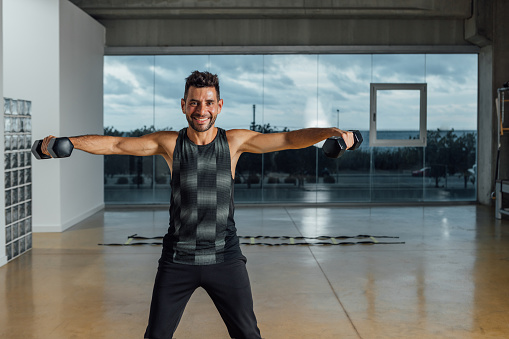 Portrait of a smiling caucasian male personal instructor, looking at camera, exercising with dumbbells in a big gym. workout lifting dumbbell weights. Horizontal, window background, Copy-space.