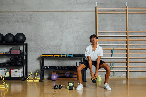 Full length Hispanic Boy Teenager exercises weight routine training with dumbbell at gym on concrete wall background, wooden bars wall. Workout, lifting weight. Copy-space, horizontal.