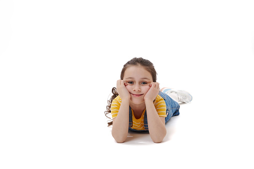 Caucasian adorable lovely 5-6 years old child, little girl in yellow t-shirt and denim sundress putting hands under her face while lying on belly, looking at camera, isolated on white background