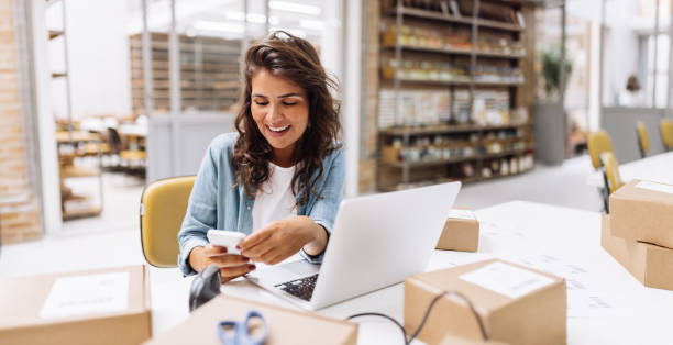 Smiling online store owner reading a text message on her smartphone Smiling online store owner reading a text message on her smartphone. Happy businesswoman making plans for product shipping in her warehouse. Female entrepreneur running an e-commerce small business. small business stock pictures, royalty-free photos & images