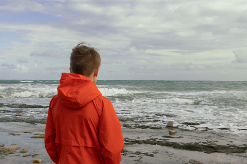 a boy with blond hair in an orange windbreaker, stands with his back on the embankment close-up, there is a place for an inscription