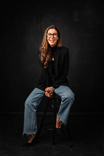 Full length of an attractive woman toothy smiling and looking at camera. Brunette haired female wearing blazer and eyewear while sitting against isolated black background. Copy space.