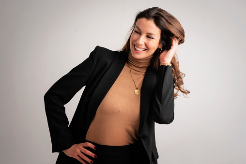 Studio portrait of an attractive woman toothy smiling and looking at camera. Brunette haired female wearing blazer and standing against isolated white background. Copy space.