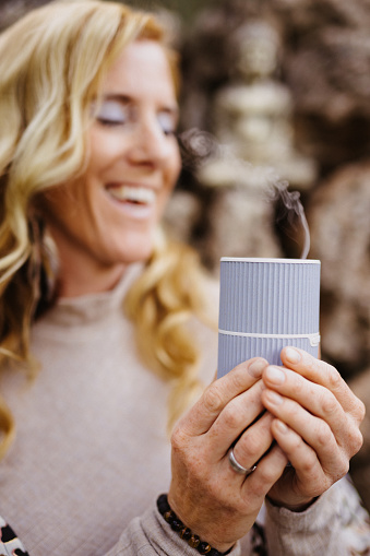 Smiling woman holding pleasurable an oil essence diffuser in front of a stone wall with a small Buddha statue. Useful for healthy, mindfulness, consciousness, awareness and self love concepts. Very selective focus and added grain. Part of a series.