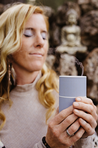 Blonde woman holding pleasurable an oil essence diffuser in front of a stone wall with a small Buddha statue. Useful for healthy, mindfulness, consciousness, awareness and self love concepts. Very selective focus and added grain. Part of a series.