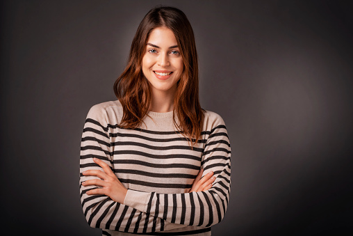 Portrait of smiling woman with arms crossed. Confident young female is having brunette hair. She is in striped  sweater against dark background. Copy space.