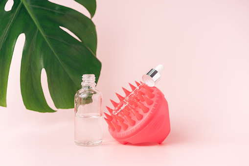 Massager for scalp and cosmetics against pink background and monstera leaf.