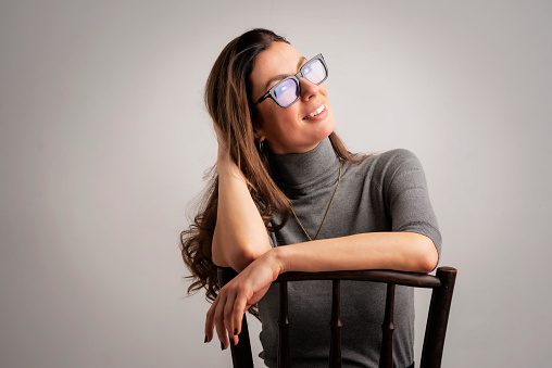 Close-up of an attractive middle aged woman with toothy smile wearing turtleneck sweater and eyewear while sitting at isolated grey background. Copy space. Studio shot. Hand on head.