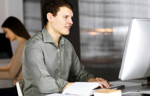 istock Dark-haired young businessman and programmer in a green shirt is working hard on his computer, while sitting at the desk in a modern cabinet with a female colleague on the background. Concept of successful business start up 1483318165