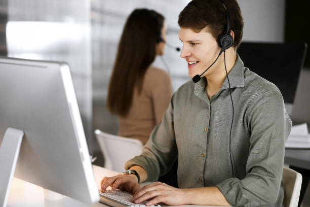 istock Young dark-haired guy in a green shirt and headsets is talking to a client, while sitting at the desk, working together with a female colleague in a modern office. Call center operators at work 1483318187