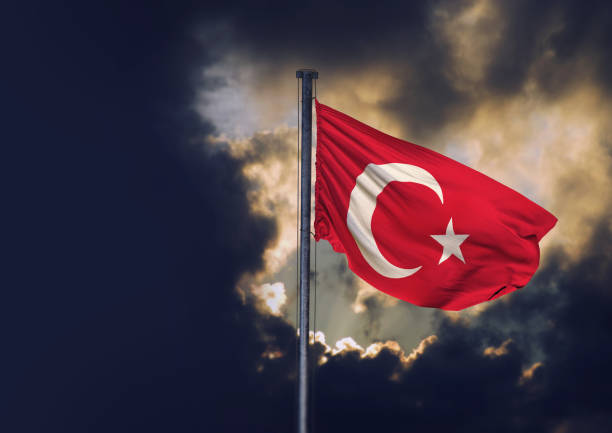 A red warning flag flutters in the wind against a clear sky with the sun low on the horizon, casting a glow around the flagpole. A lone bird glides across the open sky, adding a sense of stillness and vastness to the scene.