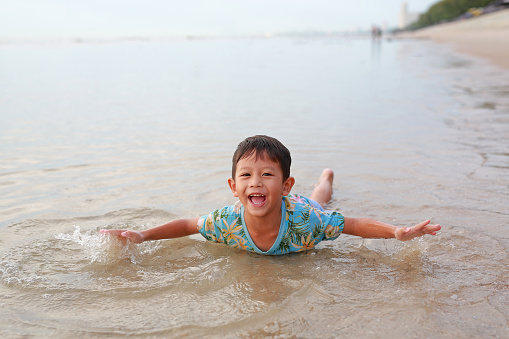 Cheerful Asian boy enjoy playing and lying on tropical sand beach at sunrise. Adorable little child kid having fun in summer holiday.