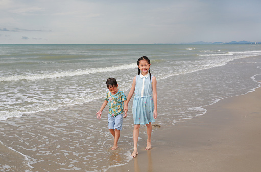 Happy Asian young sister and little brother walking together on tropical sand beach at sunrise. Happy family boy and girl enjoy in summer holiday.