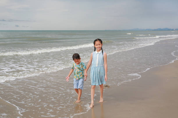 Happy Asian young sister and little brother walking together on tropical sand beach at sunrise. Happy family boy and girl enjoy in summer holiday. Happy Asian young sister and little brother walking together on tropical sand beach at sunrise. Happy family boy and girl enjoy in summer holiday. family beach vacations travel stock pictures, royalty-free photos & images