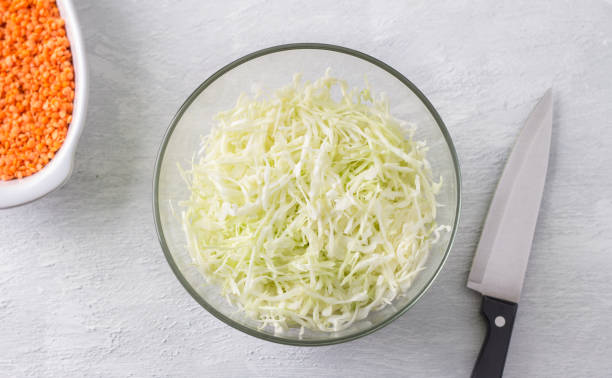 un tazón de vidrio de repollo picado, una fuente para hornear con lentejas rojas y un cuchillo sobre un fondo gris claro. cocinar comida vegana casera - sauerkraut cabbage vegetable white cabbage fotografías e imágenes de stock