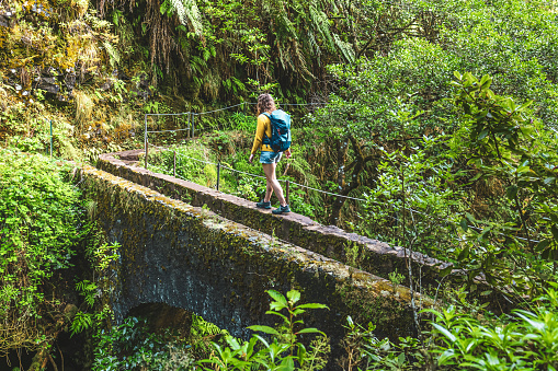 Description: Backpacker woman walking on overgrown footpath over old bridge in Madeira's rainforest in the morning. Levada of Caldeirão Verde, Madeira Island, Portugal, Europe.