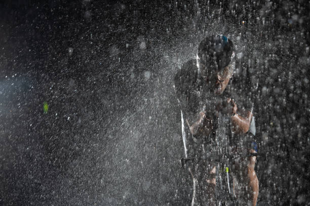 A triathlete braving the rain as he cycles through the night, preparing himself for the upcoming marathon. The blurred raindrops in the foreground and the dark, moody atmosphere in the background add to the sense of determination and grit shown by the athlete