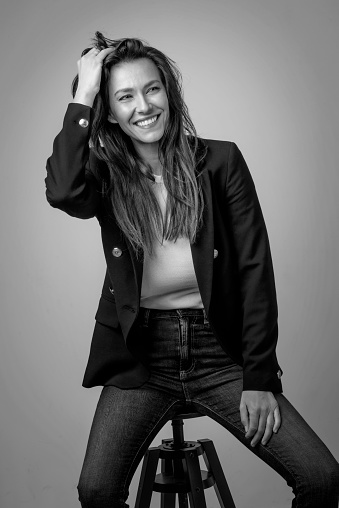 Studio portrait of attractive woman wearing blazer and jeans while sitting at isolated background. (B&W) Copy space.