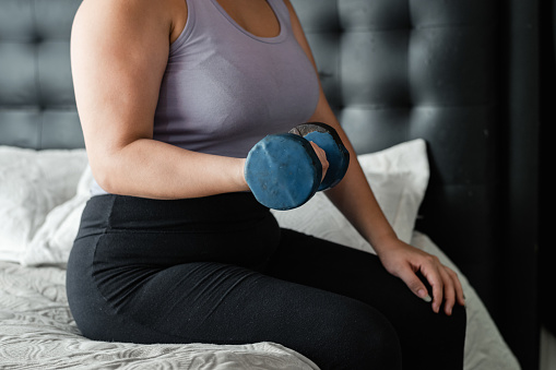 medium shot of a latina woman doing a seated exercise on her bed with a blue dumbbell. working the biceps.