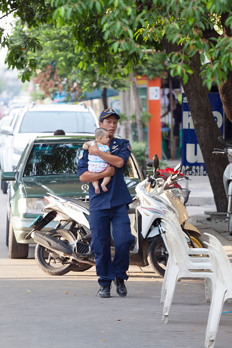 Thai man in security staff clothes with baby on arms is walking in street Chokchai 4 in Bangkok ladprao. At right side are chairs of street kitchen market stall. man is looking into camera. In right background is a tree