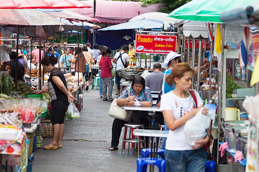 Scene at local street food and fruit market in Bangkok Ladprao. Market is located in street Chokchai 4. View along market stalls at left side and street kitchens at right side. People are shopping. In center a woman is sitting and eating.