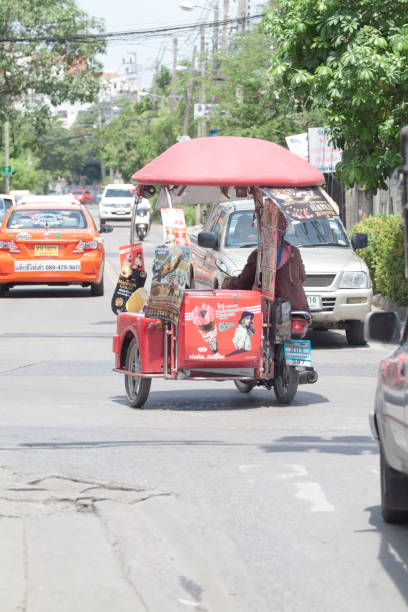 Thai ice cream vendor on motorcycle with carrier is driving in street in Bangkok Ladprao. Person is selling Langnese aka asian Wallis ice cream brand. At motorcycle is a red umbrella as uv protection. In street cars are parked and a taxi is driving in background