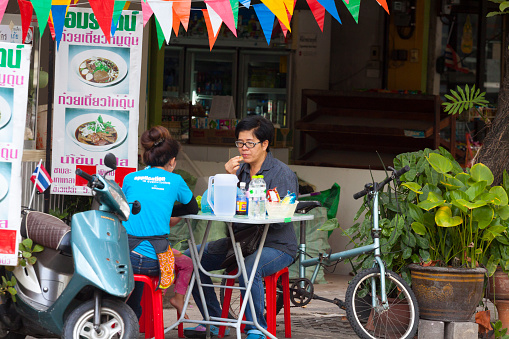 Two adult thai women have breakfast in street in front of shop in Nathon on islkand Ko Samui in south of Thailand. Shop is decorated withy rainbow colored penants. One woman is wearing glasses