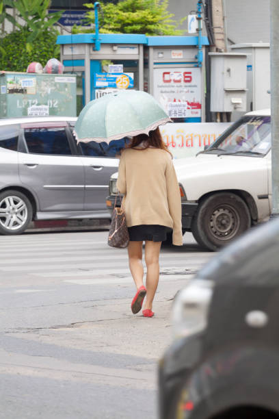 Thai woman with umbrella as uv protection is walking over street Chockchai 4 in Bangkok Ladprao. Woman is wearing a skirt and has a bag. On street is some traffic