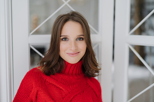 Headshot of charming woman with dark hair, wears knitted sweater, looks directly at camera, has friendly smile, poses indoor, has pleasant talk with boyfriend. People, lifestyle, spare time concept