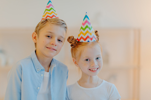 Horizontal shot of happy girl and boy wears cone party hats, celebrate birthday together, have good mood, wait for guests, pose indoor against blurred background. Children, holiday, celebration