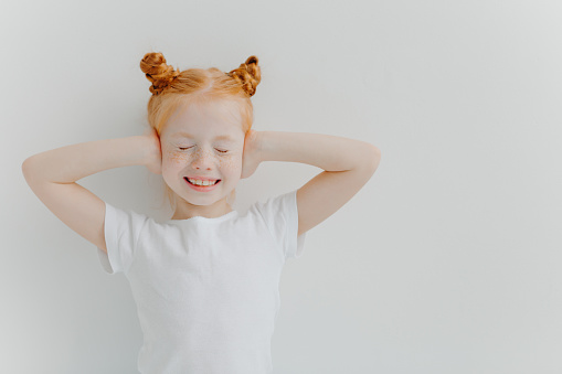 Positive good looking ginger freckled beautiful girl covers ears with hands, hears very loud music, closes eyes and smiles broadly, wears white t shirt, stands indoor, being on children party